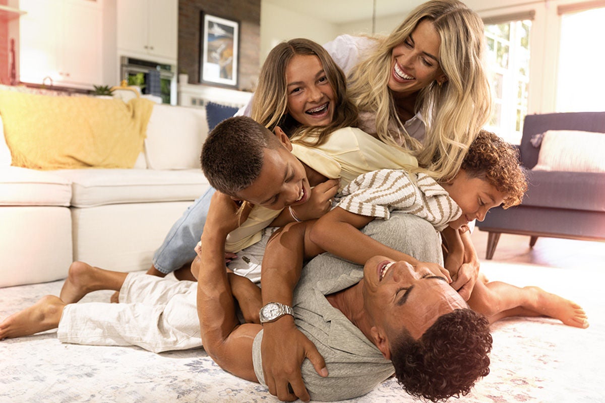 A joyful family piles on top of a man lying on the floor, laughing and playing together. They are in a bright, modern living room with a white sofa and soft natural light. Two children are on top of the adults, smiling and hugging. The scene conveys warmth, affection, and energetic fun in a cozy home environment.