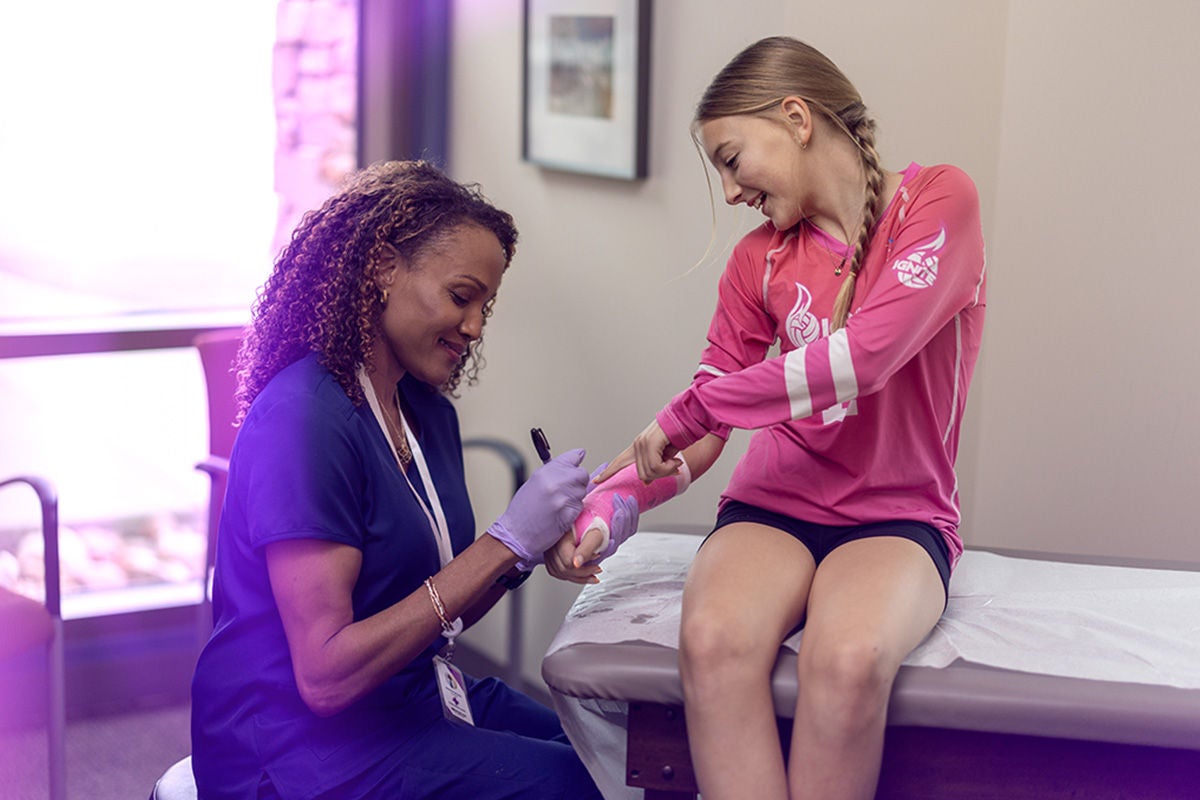 A healthcare professional in scrubs attends to a teenage girl sitting on an examination table. The nurse is writing or drawing on the girl's pink arm cast while wearing medical gloves. The setting appears to be a clinic or doctor's office with neutral walls and framed artwork. The girl smiles and looks at her cast, suggesting a friendly and reassuring medical visit.