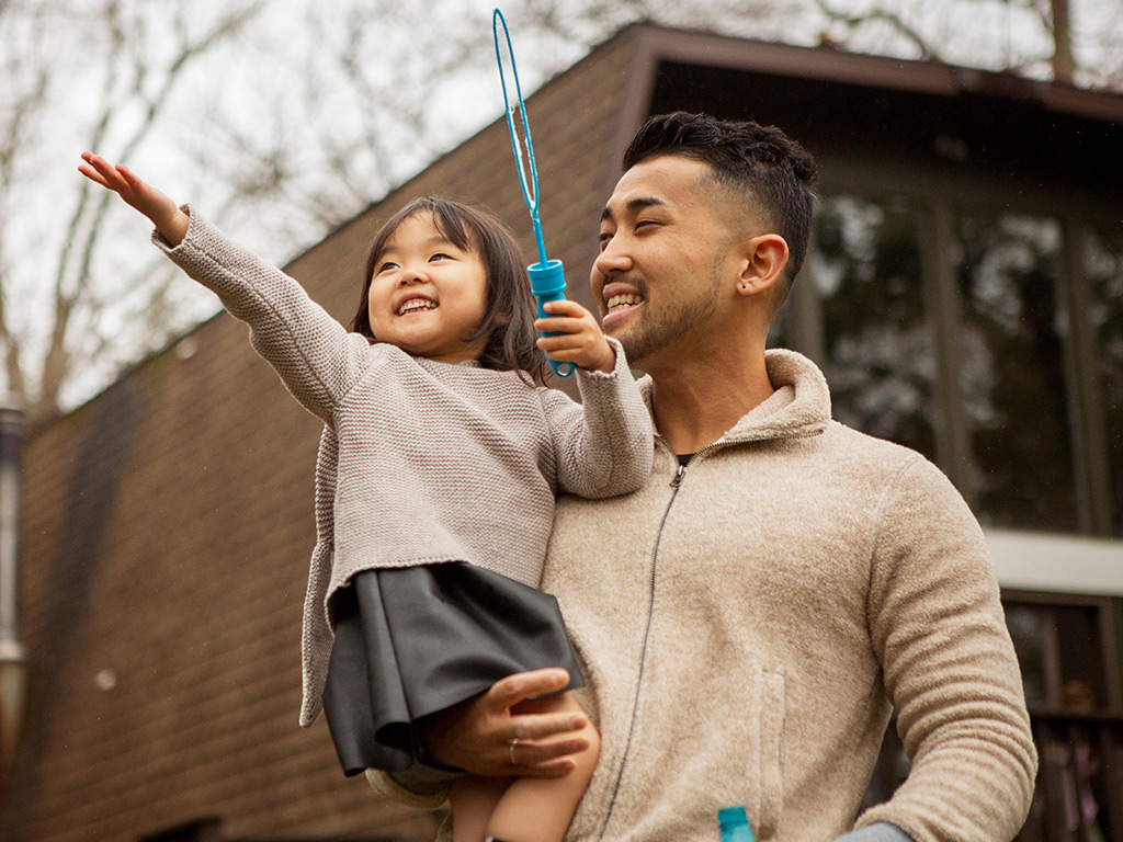 A smiling man holds a young girl in his arms as she reaches upward with a bubble wand. They are outdoors in front of a house with brown siding and bare trees in the background. Both wear neutral-toned sweaters, creating a warm and cozy feel. The scene captures a joyful family moment and playful interaction.