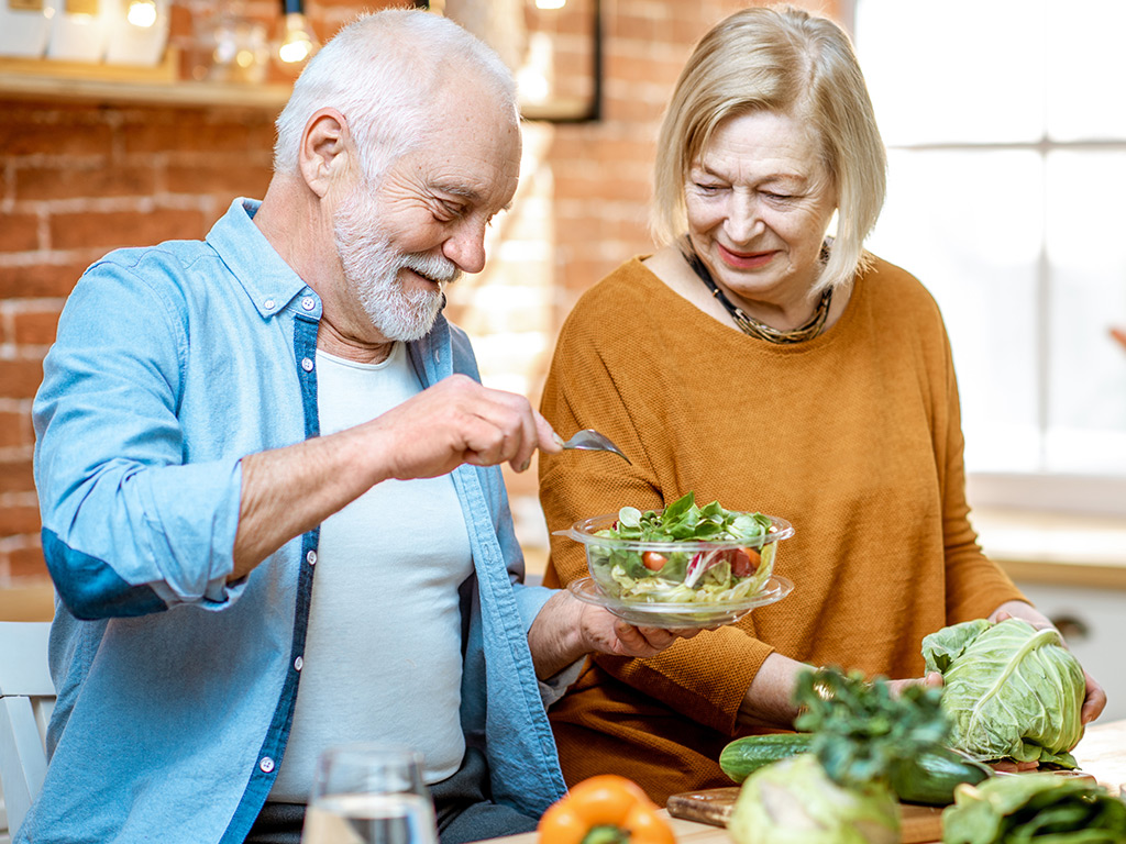 an older couple makes a green salad in a kitchen.