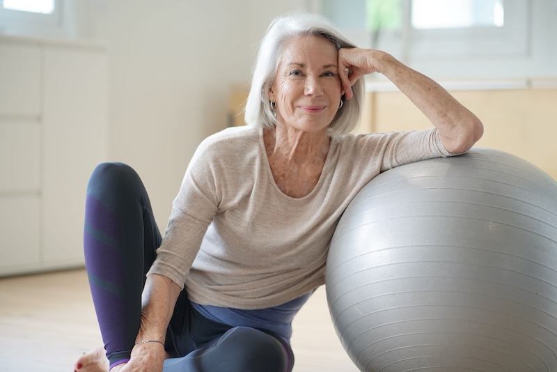  Smiling elderly woman resting on a swiss ball at home                              