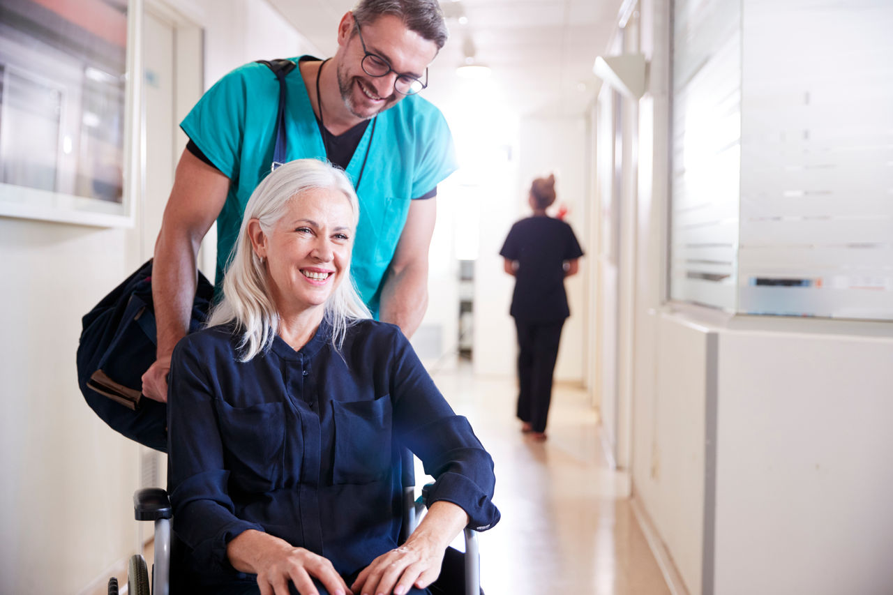 Male Orderly Pushing Senior Female Patient Being Discharged From Hospital In Wheelchair