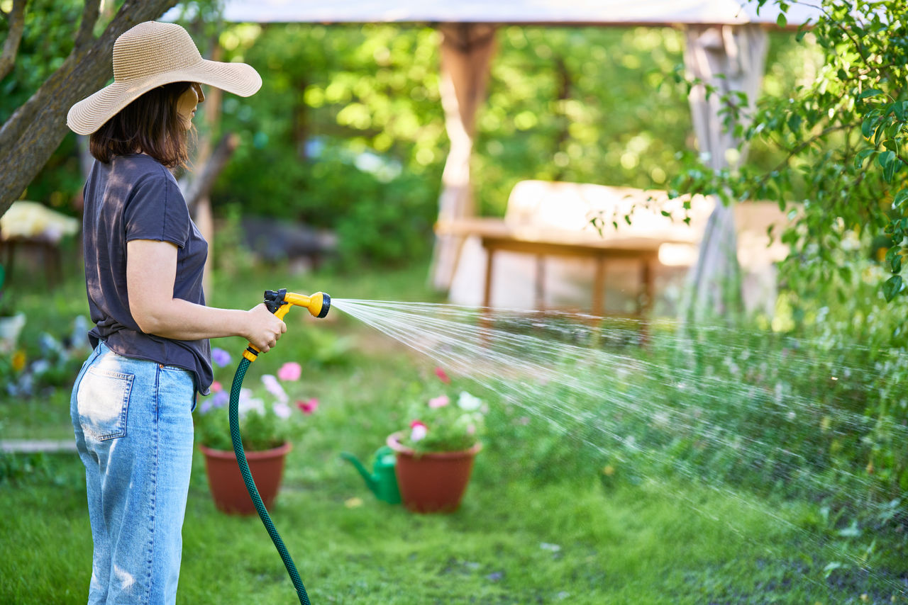 Young woman in hat watering flowers and plants in garden with hose in sunny blooming backyard. Gardening work, summer time