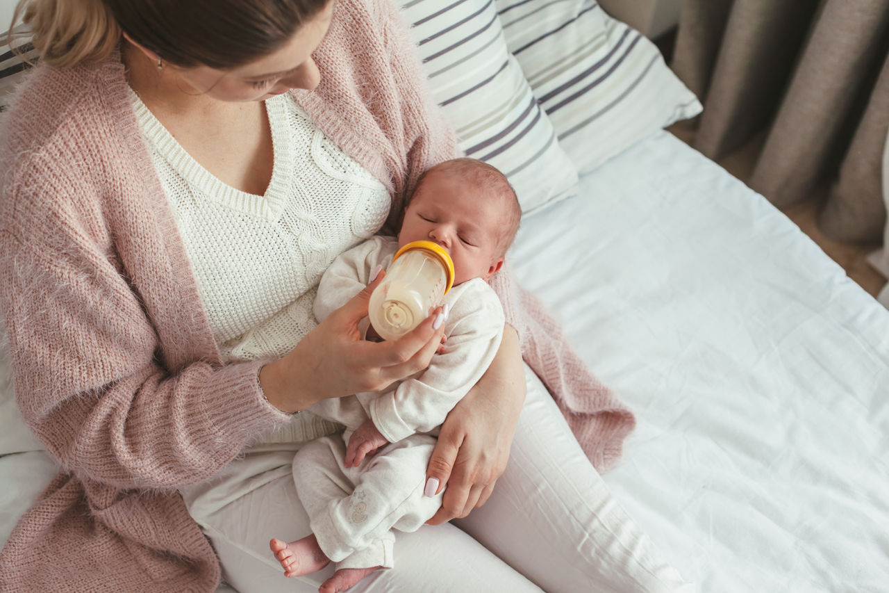 Home portrait of a newborn baby with mother on the bed. Young mom feeding her child from bottle.