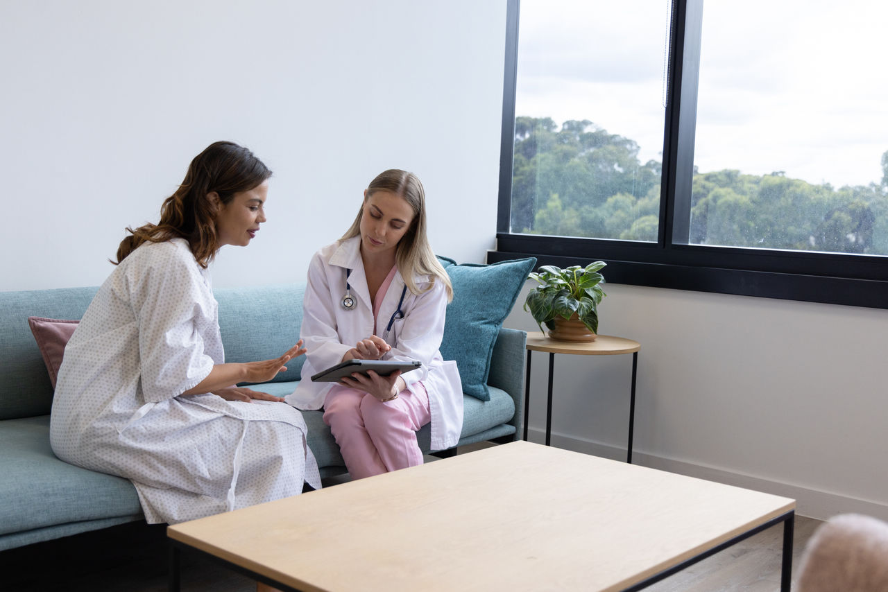 A female doctor in a white coat sits beside a patient wearing a hospital gown on a teal sofa. They are engaged in a medical consultation while looking at a digital tablet together. The scene takes place in a bright, modern clinic room with large windows and a potted plant. Natural light and minimalist furniture create a calm, professional atmosphere.