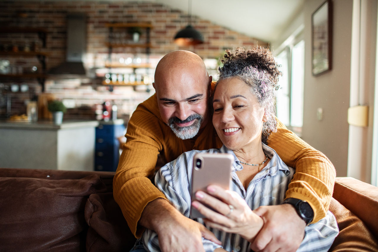 Mature couple hugging and smiling at smartphone at home