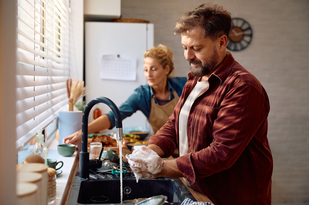 Smiling man washing dishes while cooking with his wife in the kitchen. 