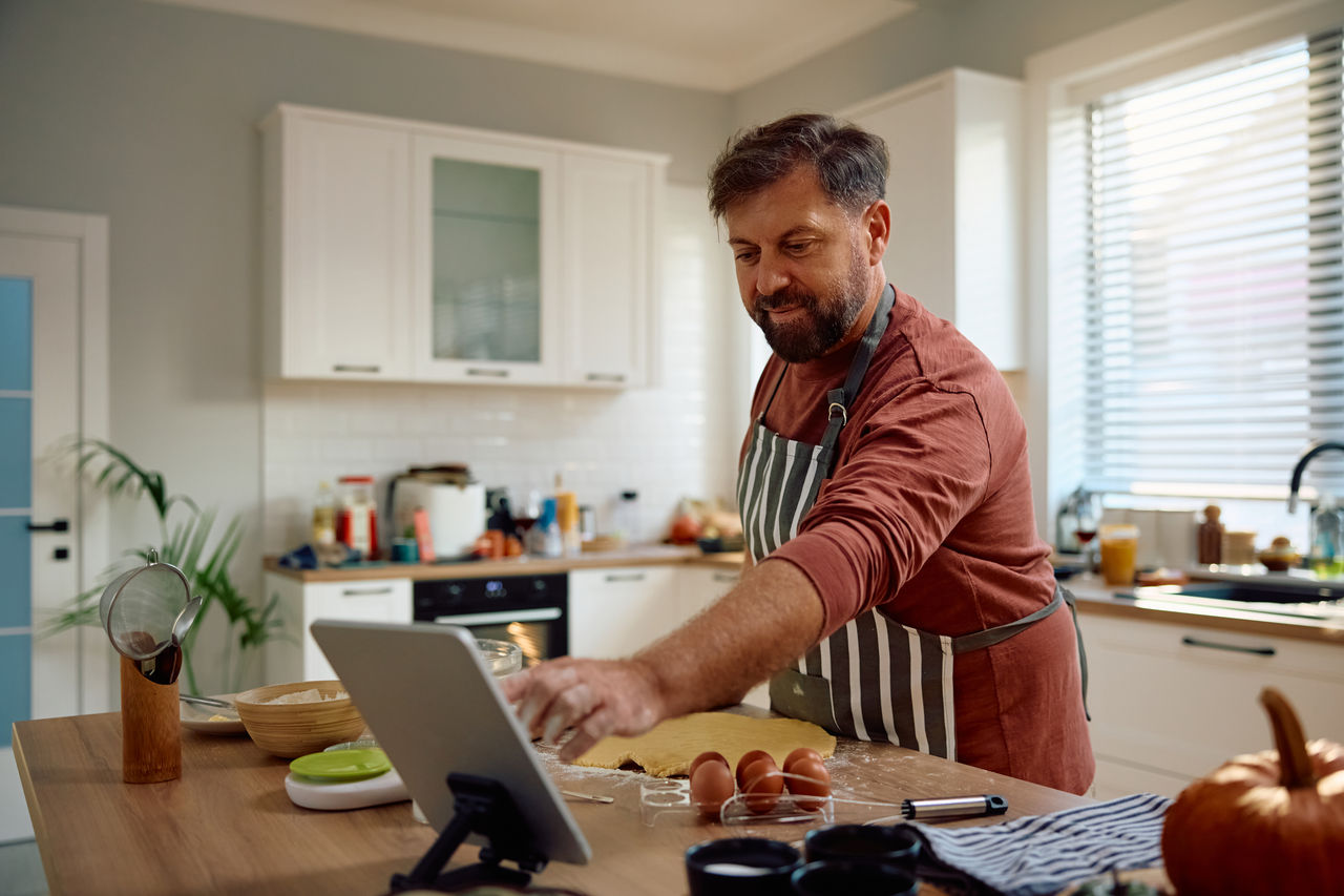 Smiling man following a recipe on touchpad while making a cake in the kitchen. 