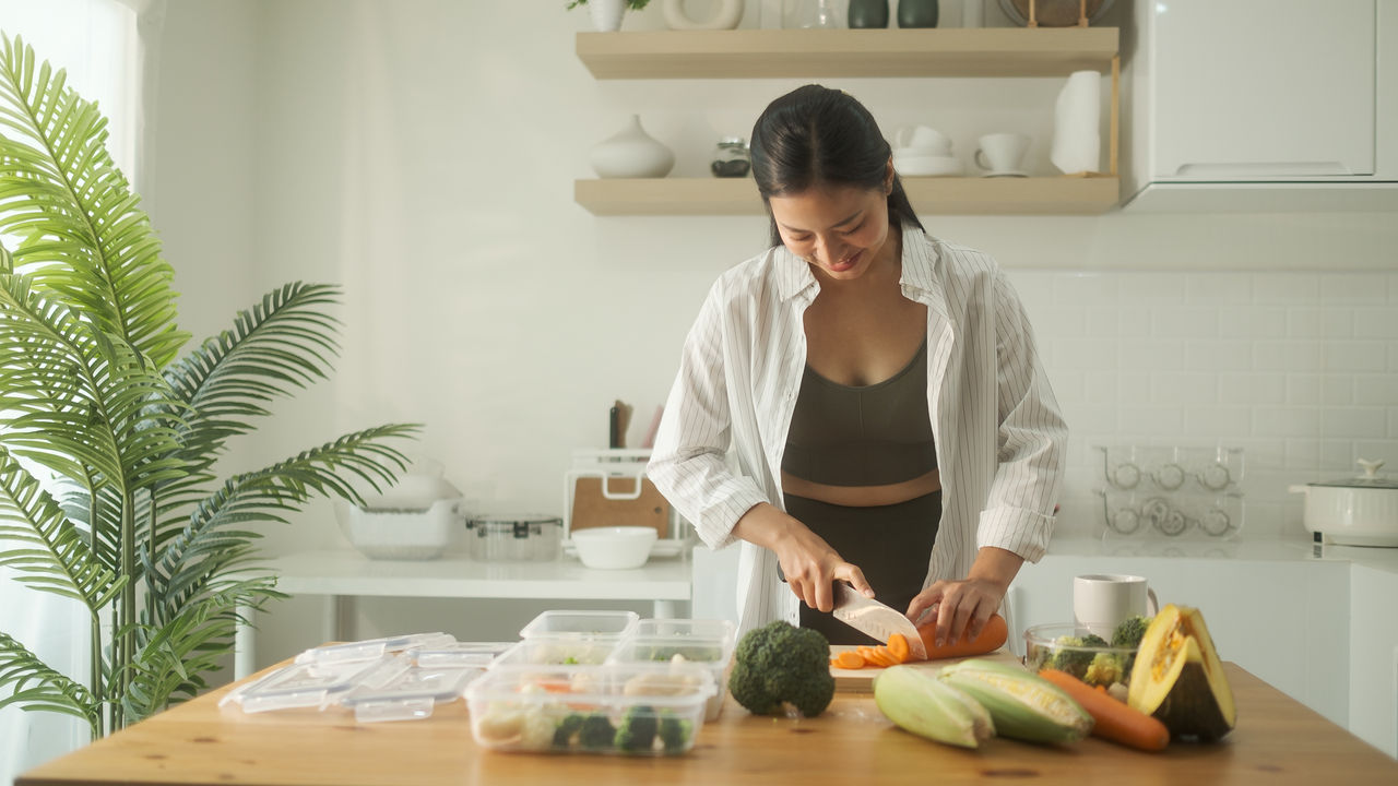 Woman dressed in fitness wear arranging meal prep containers with healthy ingredients on a wooden countertop.
