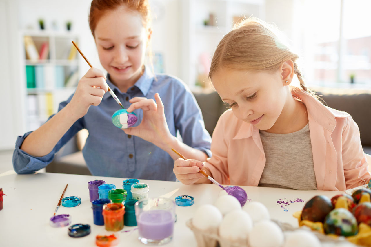 Portrait of two happy girls panting eggs for Easter in art class, lit by sunlight