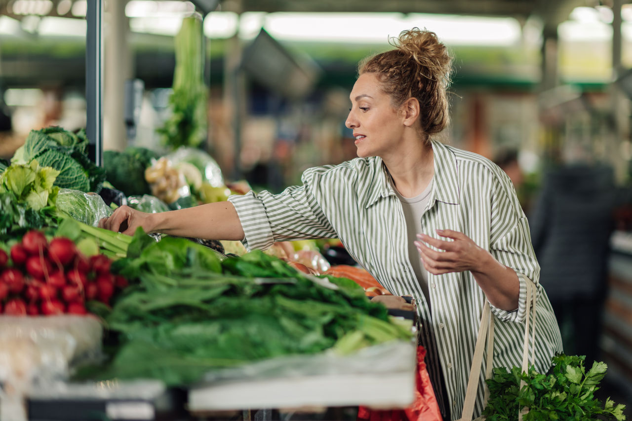 Portrait of smiling female customer standing near market stall at farmer's market and buying fresh organic vegetables. Happy shopper woman choosing healthy vegetables from organic producers at market
