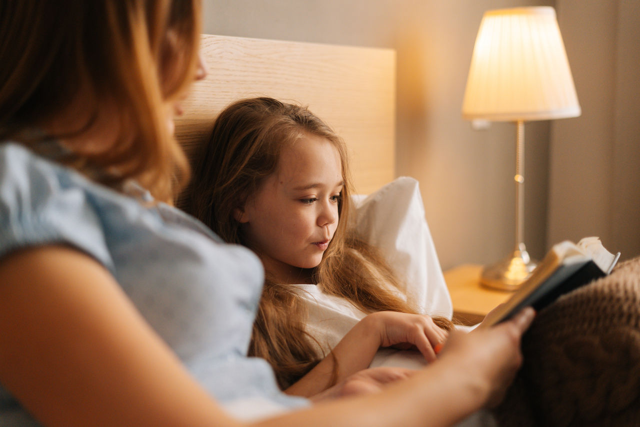 Close-up portrait of happy young mother and adorable daughter reading together children book before going to sleep while lying in bed in dark nursery bedroom, near lamp.