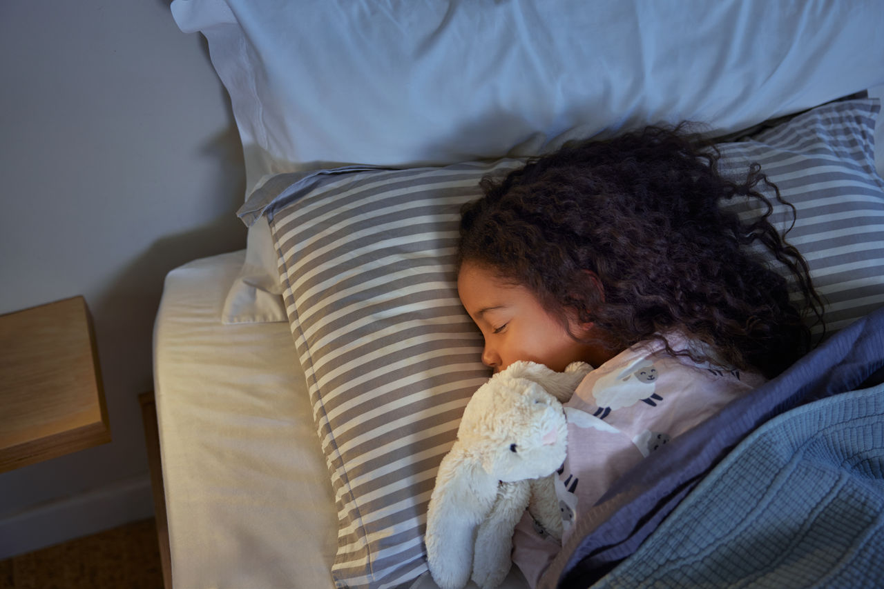 Mixed race little girl sleeping peacefully in bed, cuddling a soft toy. A serene moment of an indian female child resting comfortably in a cozy bed with the bedside lamp on. Little multiethnic girl sleep, holding her stuffed bunny close in warm light.