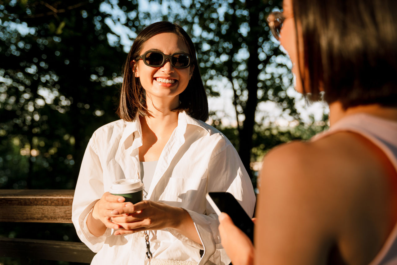 Young beautiful smiling happy woman in sun glasses looking at her friend with phone, while they standing in the sun in park