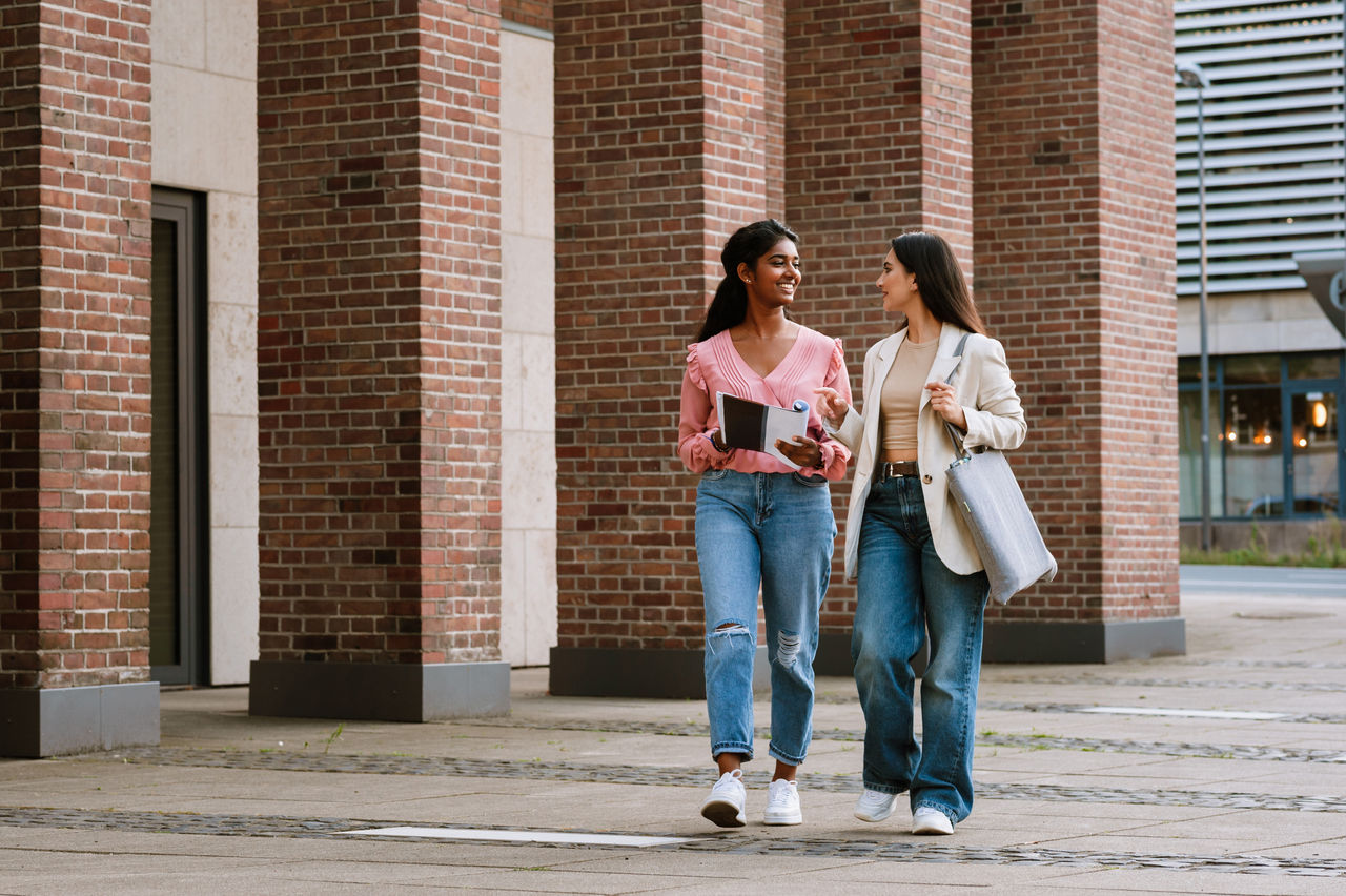 Two multinational happy women talking together while walking on city street