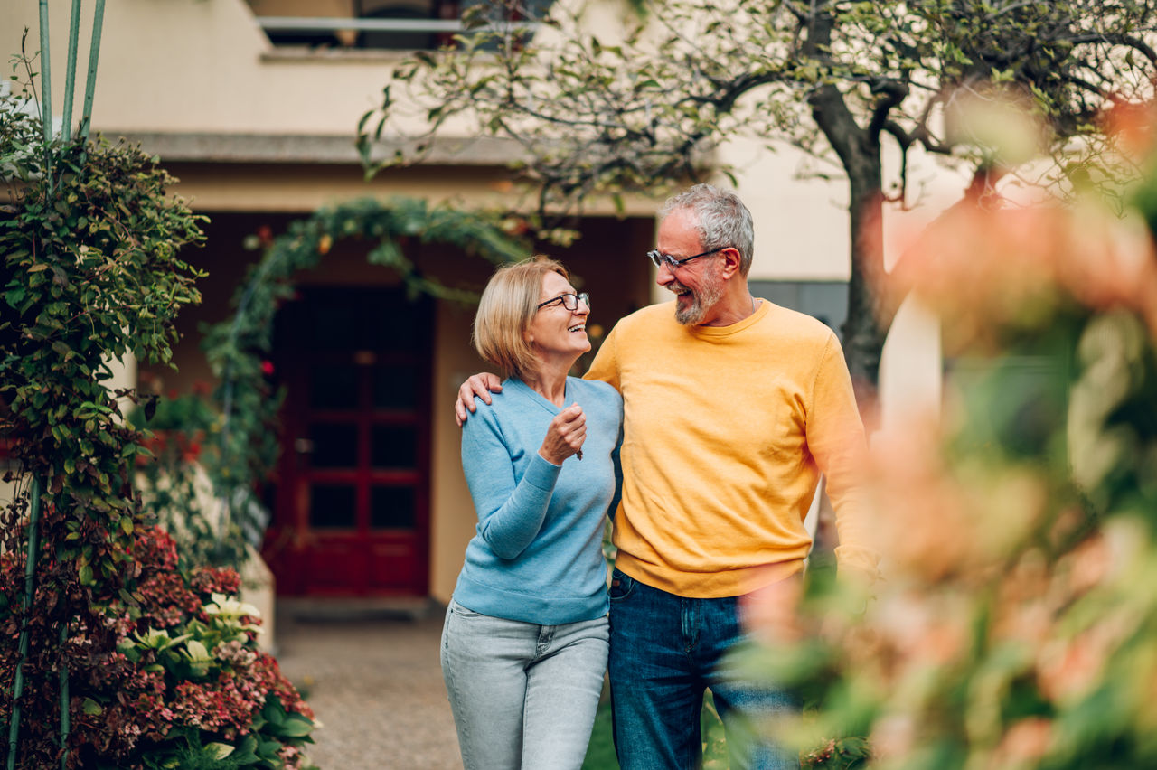 Happy mature couple bought their new home and standing in front of it and showing keys. Real estate owners. Senior married couple posing in front of their new family house. Looking at each other.