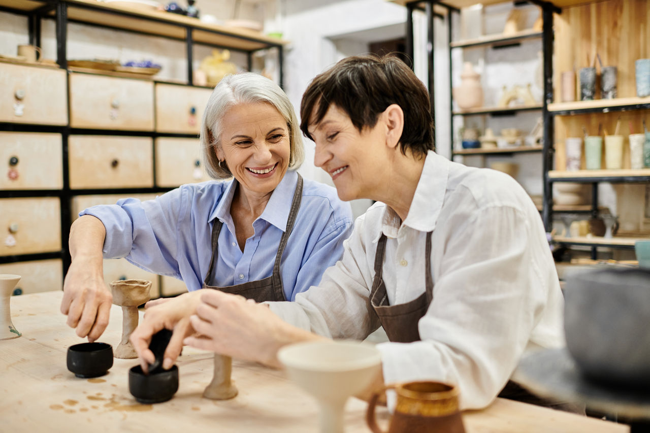 A mature lesbian couple smiles while crafting pottery in a cozy studio.