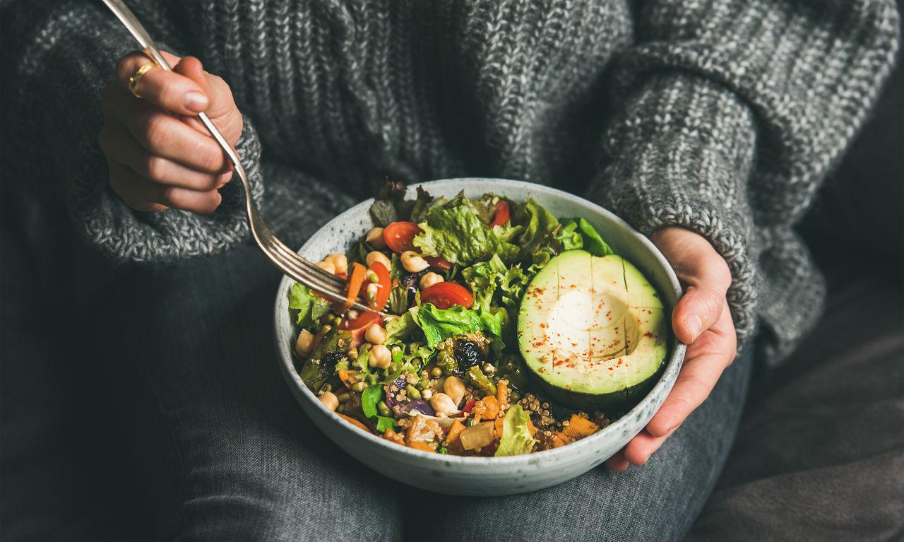 Woman eating healthy vegetarian dinner from Buddha bowl, close-up Healthy vegetarian dinner. Woman in jeans and warm sweater holding bowl with fresh salad, avocado, grains, beans, roasted vegetables, close-up. Superfood, clean eating, vegan, dieting food concept