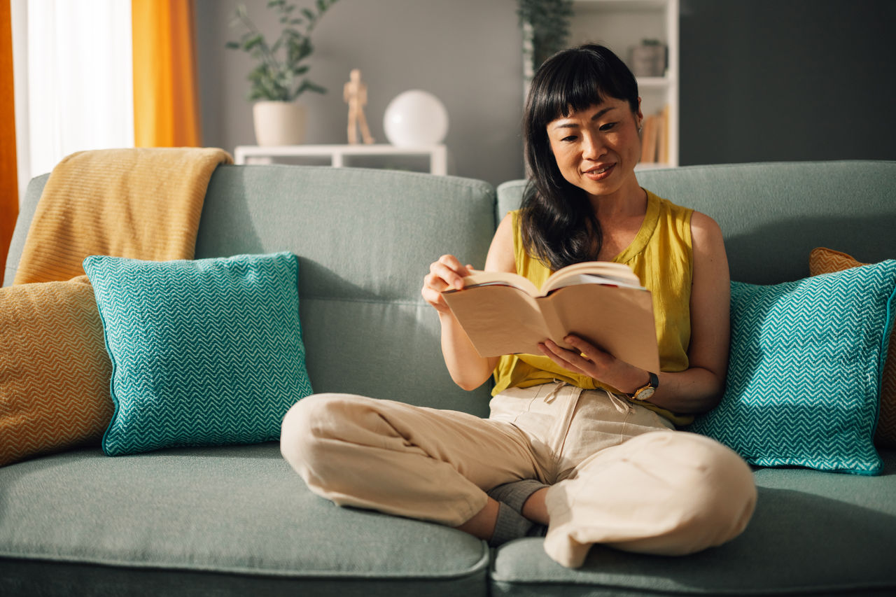 Asian woman reading comfortably on sofa with colorful cushions. Asian serene woman with a yellow top reads a book while sitting on a cozy green sofa in a well-decorated living room, showcasing her peaceful engagement with literature.